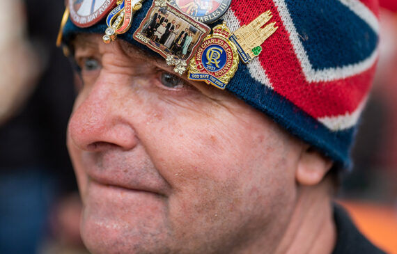 Primo piano di uomo con un cappello pieno di spille ed etichette raffiguranti i reali inglesi