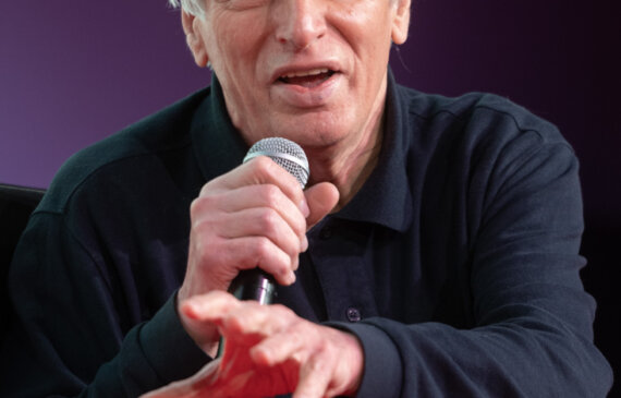 Don Luigi Ciotti sorride parlando al microfono durante una conferenza