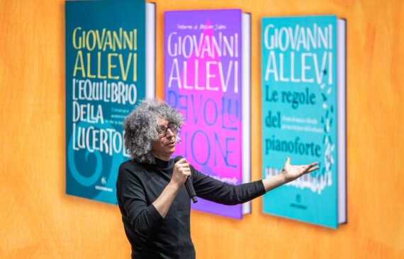 Giovanni Allevi parla al microfono su un palco con uno sfondo colorato
