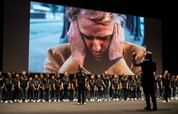 Un coro di bambini e ragazzi canta sul palco mentre sullo schermo dietro viene proiettato un fotogramma con l'attore Marlon Brando